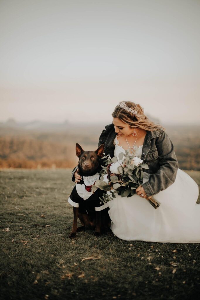 Bride in white gown wearing denim jacket holding her bouquet and petting her dog who wears a suit
