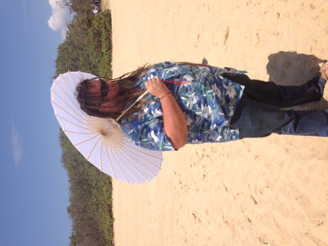 Male wedding guest with dreadlocks and sunglasses wearing tropical flowered shirt and holding a parasol on a beach 