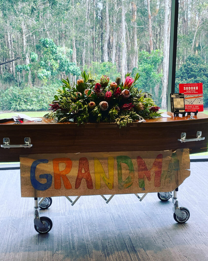 wooden casket with Australian native flowers atop and a handwritten sign saying GRANDMA at the bottom with Australian bushland in the background