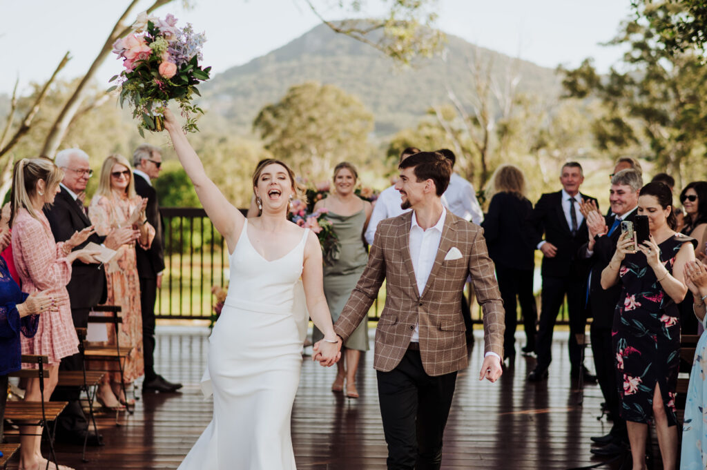 Bride with bouquet in the air, groom looking at her smiling as they walk back down the aisle as husband and wife - crowd smiling and clapping