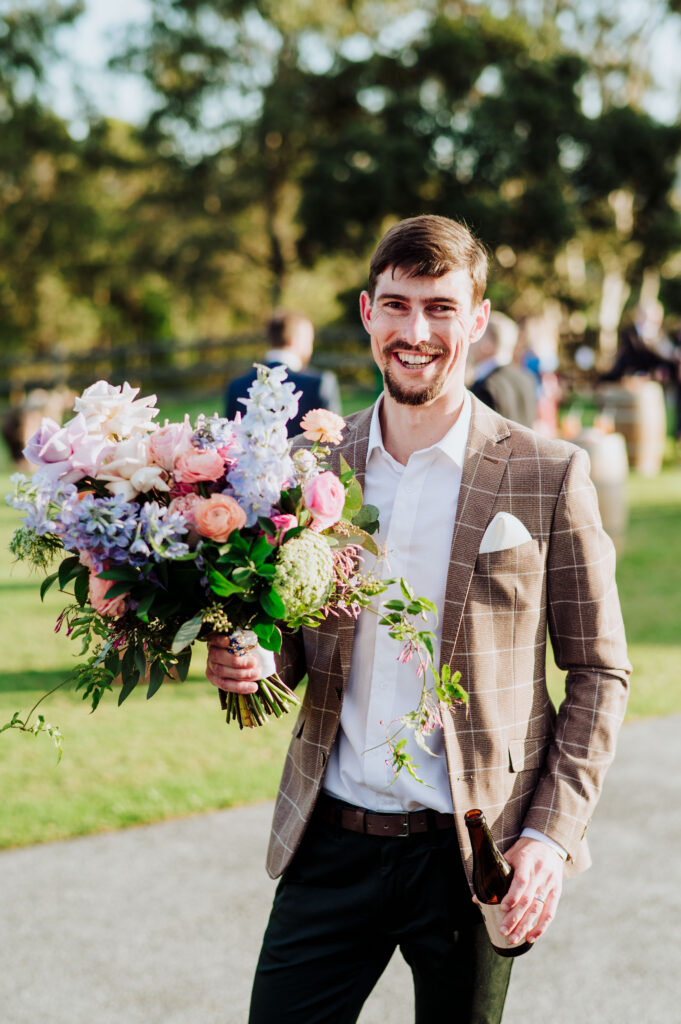 The groom in brown suit laughing as he holds the bride's colourful bouquet