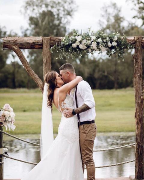 Bride and groom kissing under wedding arbour with white silk flowers and lake in the background