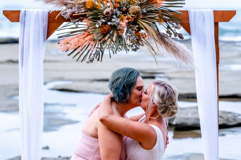 Two brides embracing and kissing under an arbour on the beach