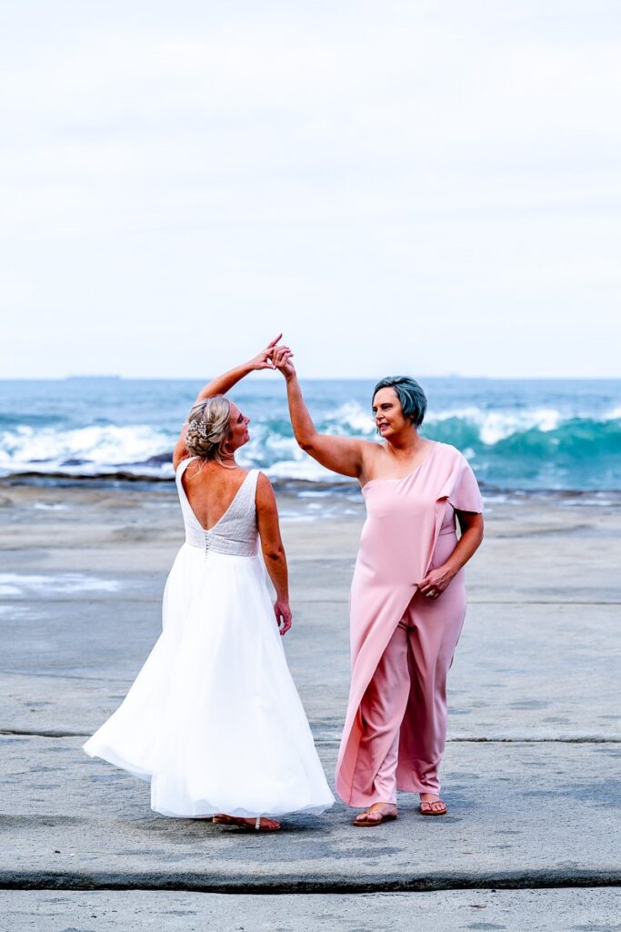 Two brides in gowns dancing on rocks overlooking a beach 