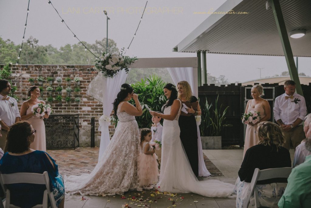 Two brides laughing as they read their vows as guests laugh and look on with marriage celebrant Lynette Maguire