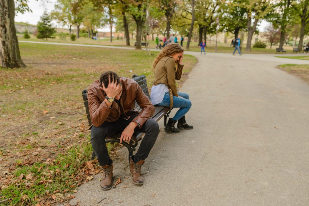 Man and woman in a park both sitting on a bench facing away from each other with hands on their heads looking sad