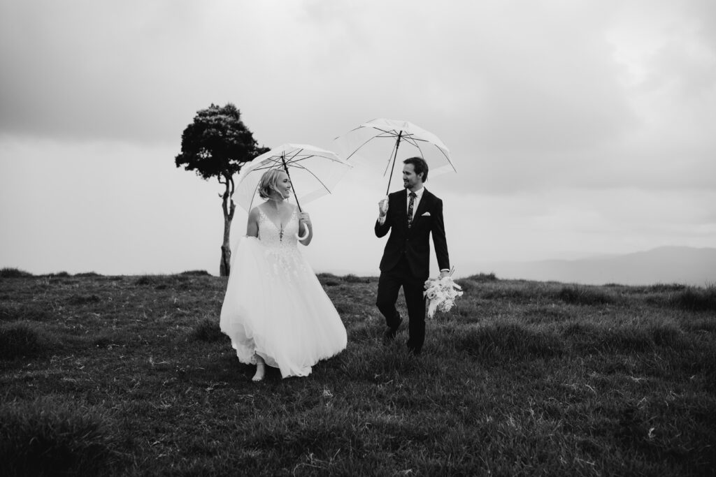 Bride and groom walking on top of a hill carrying see through umbrellas and groom carrying the bouquet while bride holds her train 