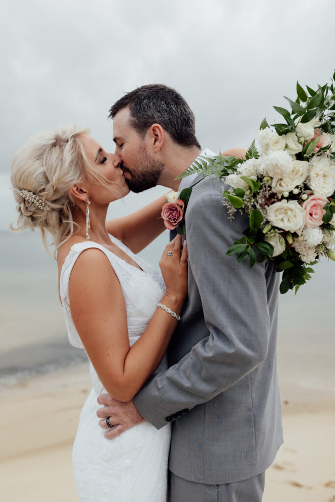 bride and groom kissing on a beach with bride holding her bouquet around his back