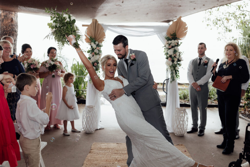 Groom dipping the bride as she holds her bouquet high in the air while laughing - crowd looking on and blowing bubbles 