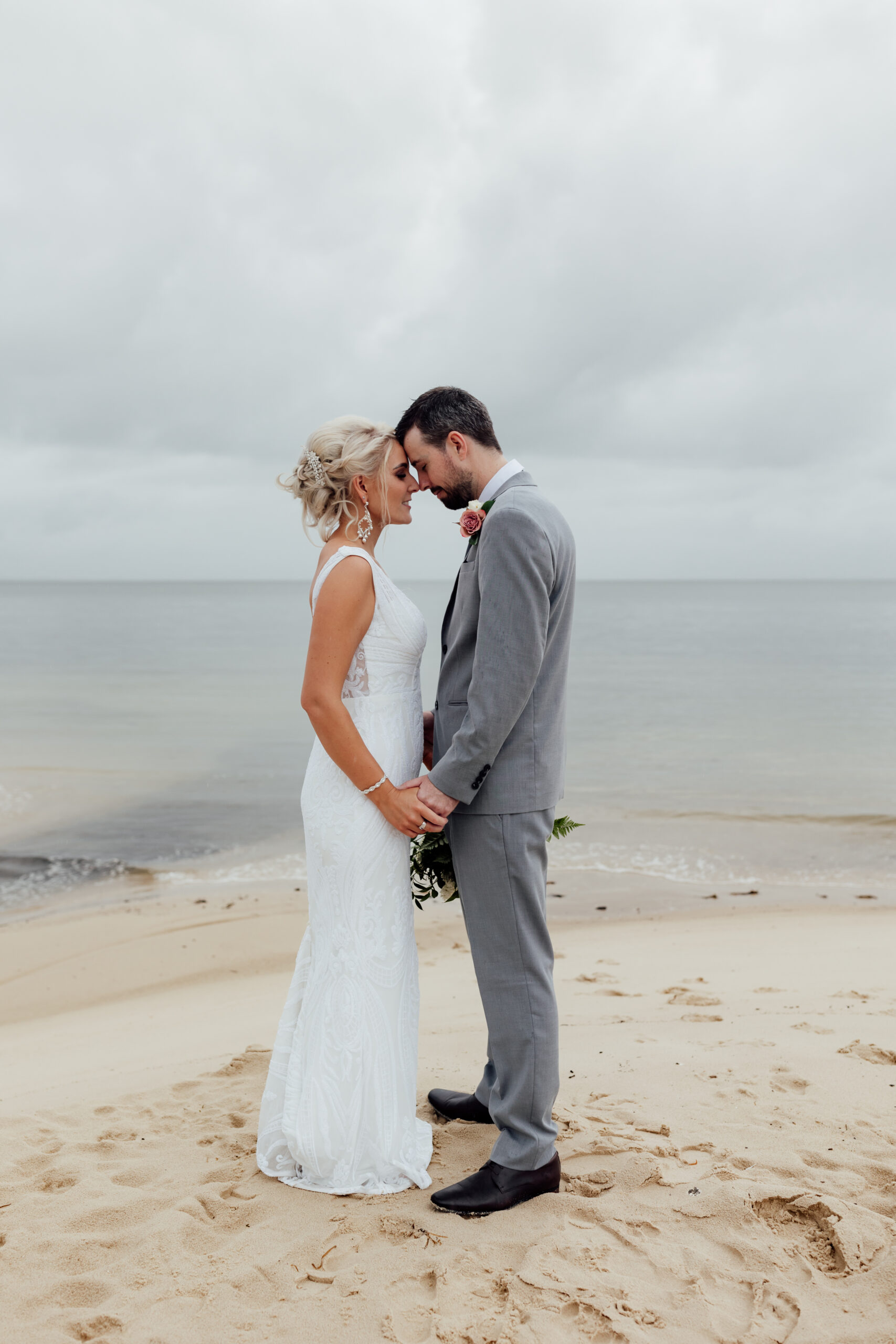 bride and groom facing each other foreheads together on beach with grey skies