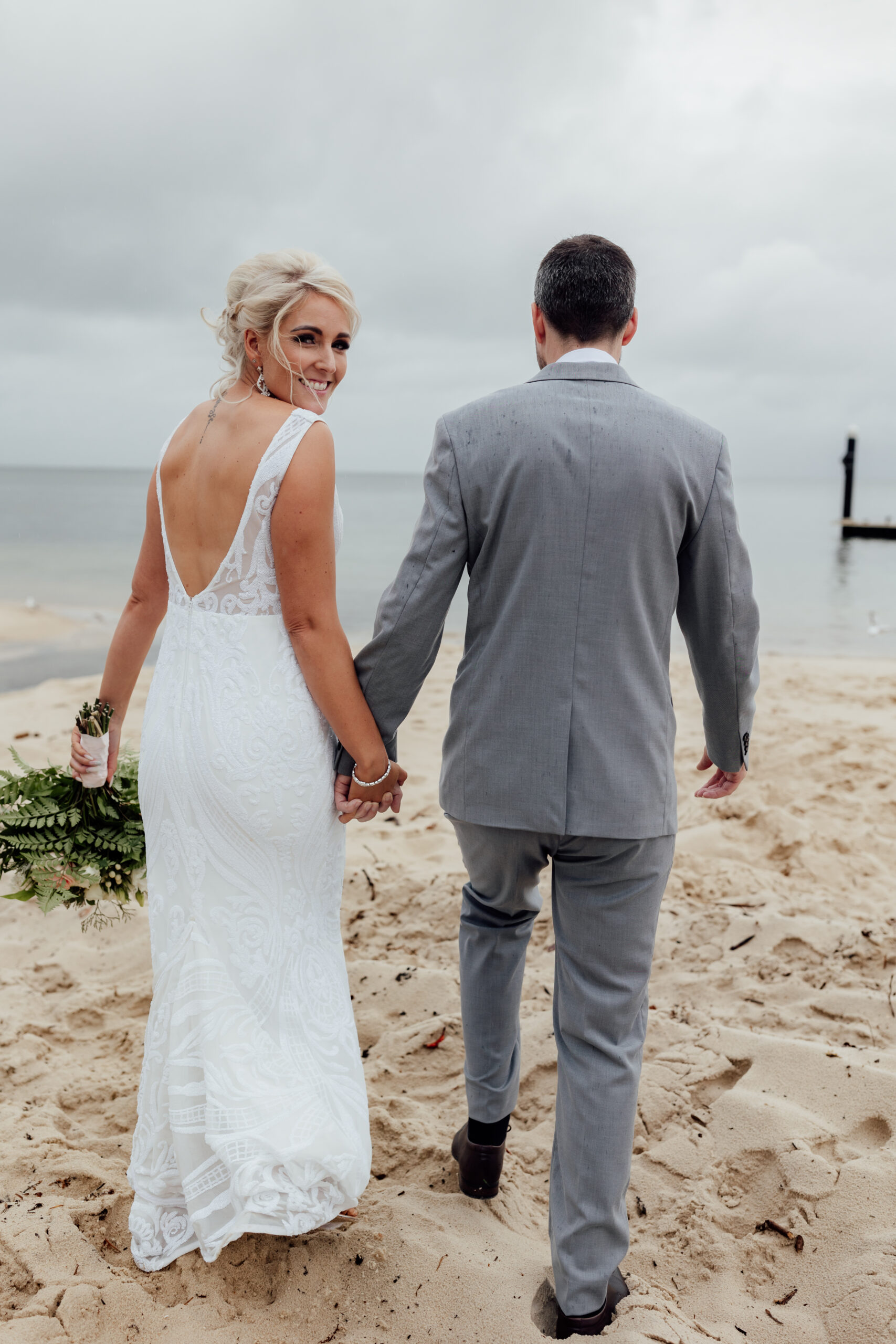 bride and groom holding hands walking on beach towards water bride smiling at camera groom's suits has rain drops with grey skies 