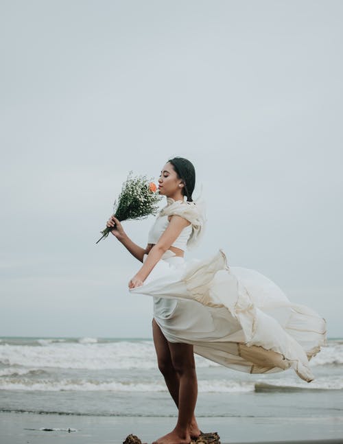 Bride on windy beach smelling her bouquet while dress blows around her 