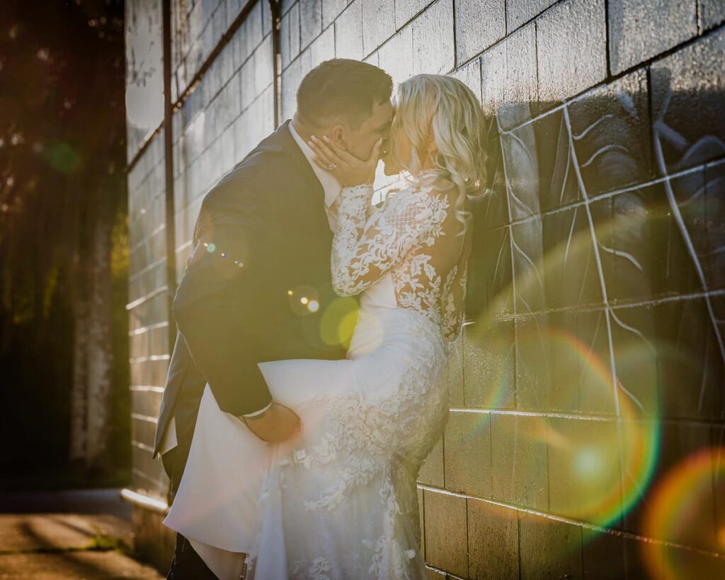 Groom kissing bride against a brick wall in the afternoon sunlight