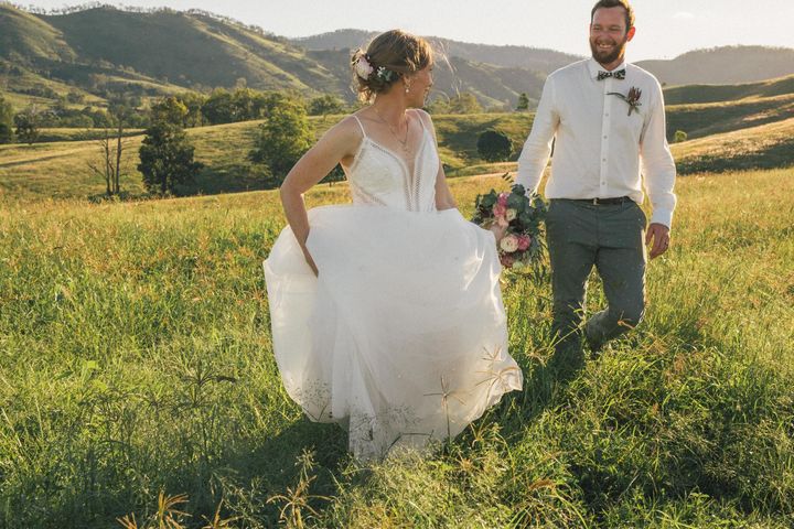 Bride and groom walking through long grassy hills, groom hold bouquet and bride hitches up her wedding dress while looking at each other and smiling 