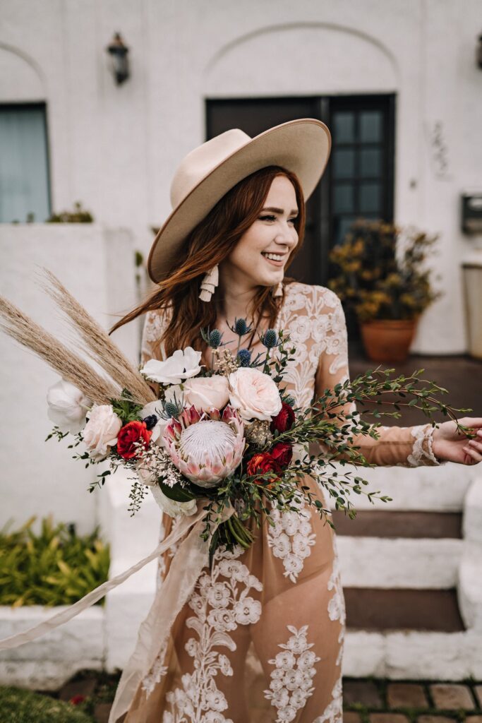 Bride wearing beige dress with white lace inserts, smiling, wearing wide brimmed had and holding big bouquet with pale pink proteas and pampas grass