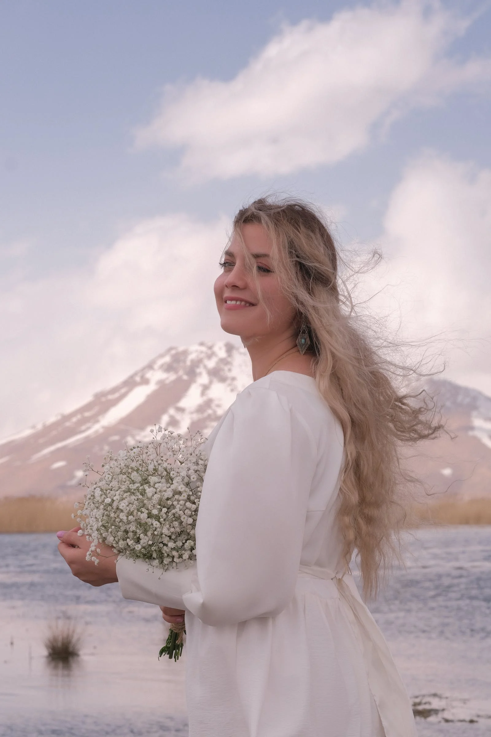 Smiling bride holding a bunch of baby's breath flowers, blonde hair tousled by the breeze, lake and mountain in the background