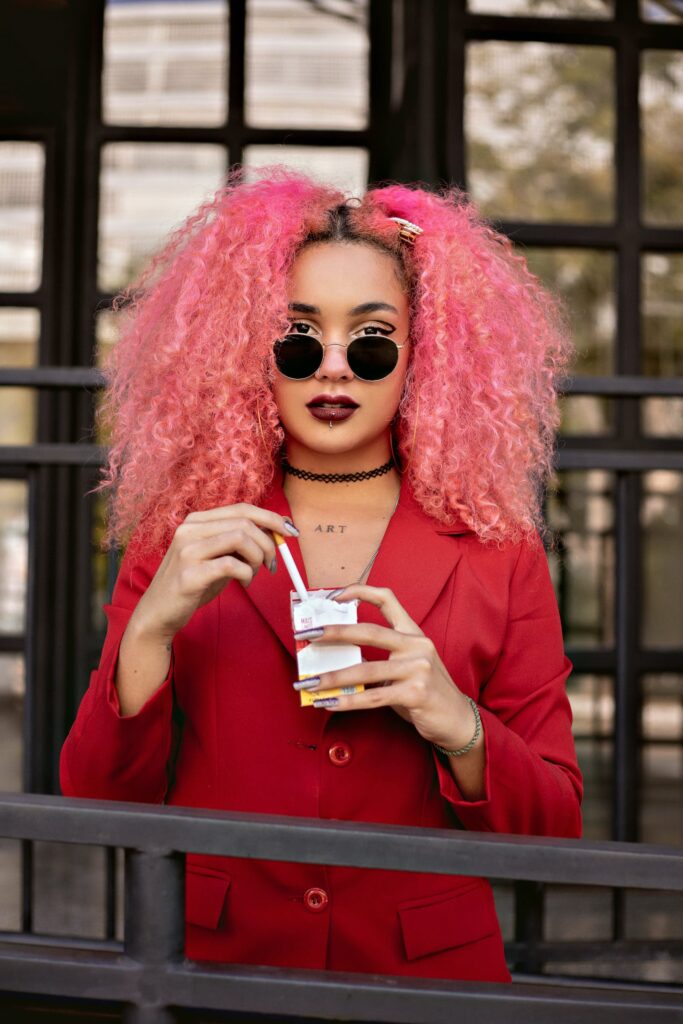 Lady with pink curly hair looks at camera while hold a drink, her round sunglasses pushed down her nose