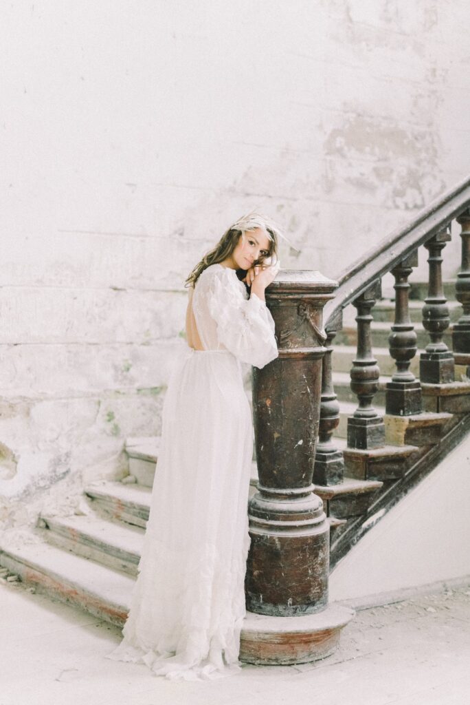 Bride in long white gown and fastenator, leaning on concrete stair pillar 
