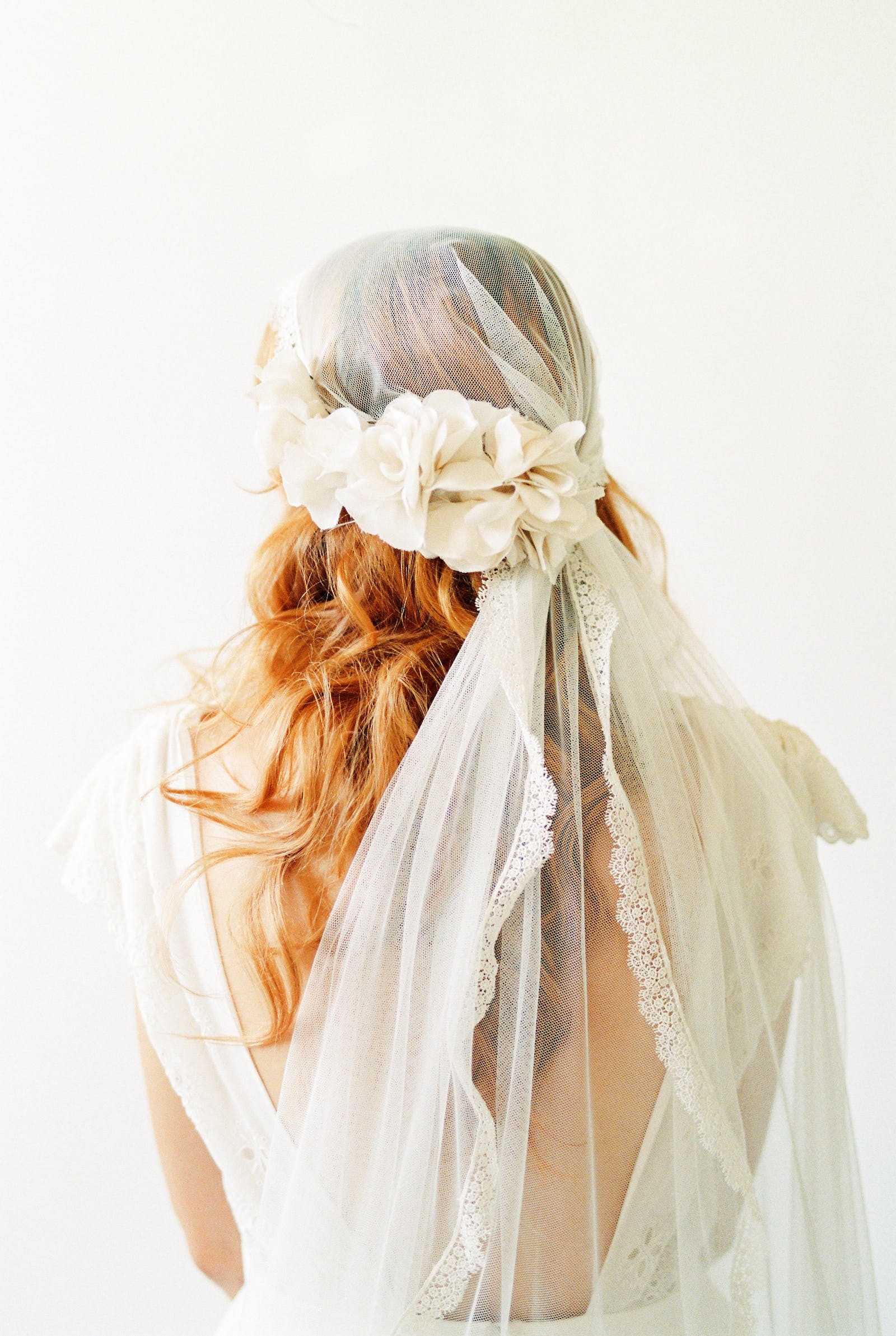 back of bride's head with 30s style veil with lace detail brought together with white silk flowers over long auburn hair  