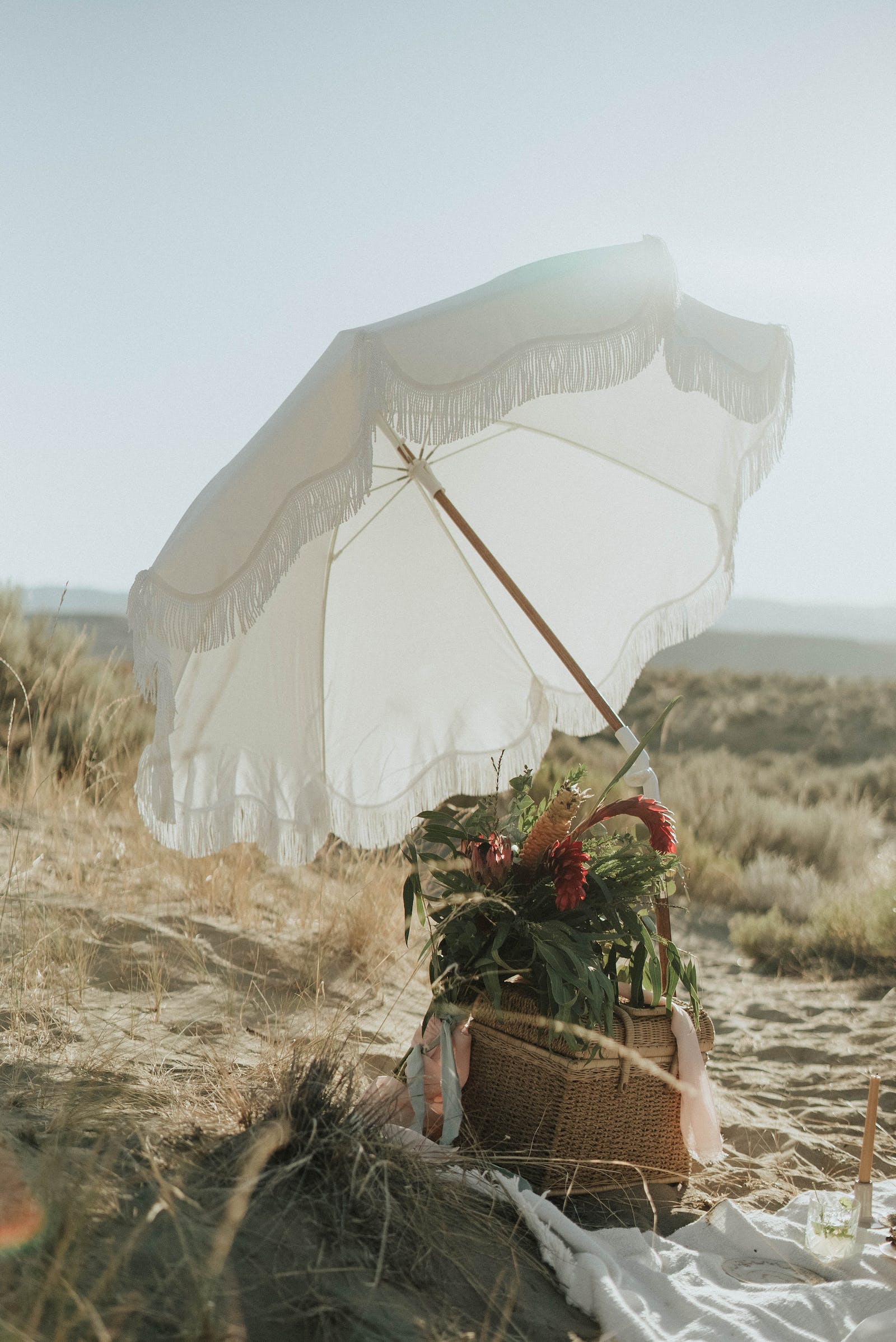 large white cloth parasol with tassles atop a picnic box and flowers resting in the dunes of a beach