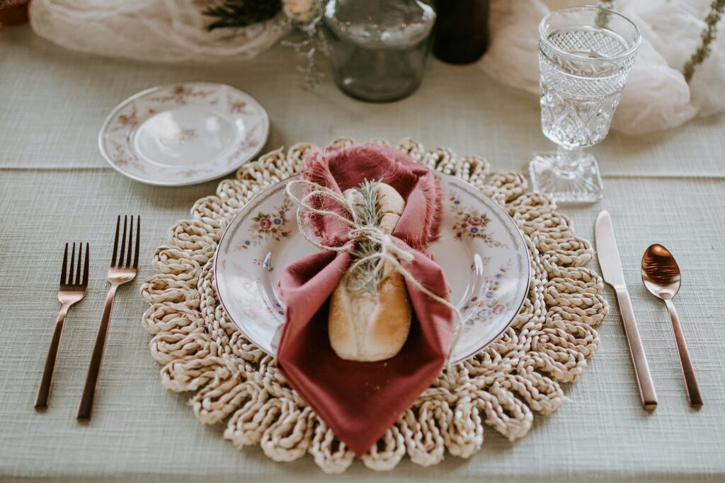 Breadroll wrapped in a russet coloured napkin tied with string sits on bread plate with flower details, which sits on round rattan placemat