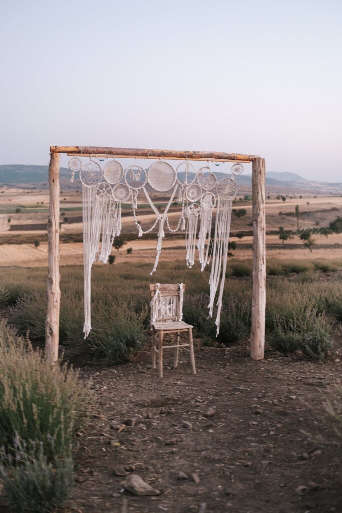 Simple arbour with one chair both decorated with macrame and set in a lavendar farm with dirt in the foreground