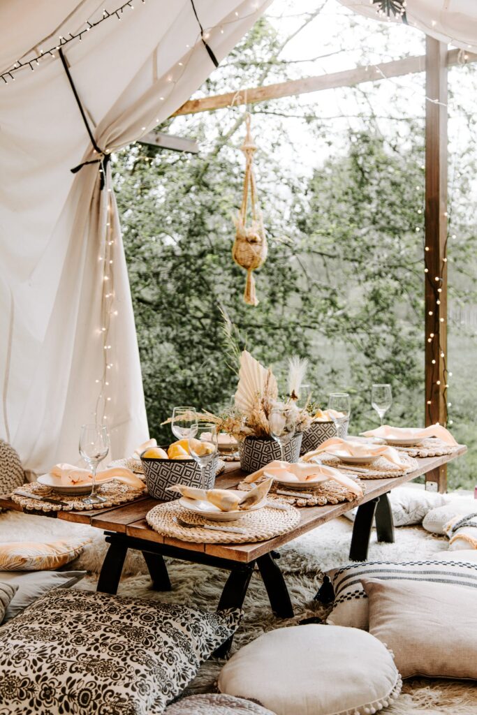 Picnic setting  with natural fabrics and cushions, set with rattan placements and bowls of fruit with dried flower setting with tree in the background