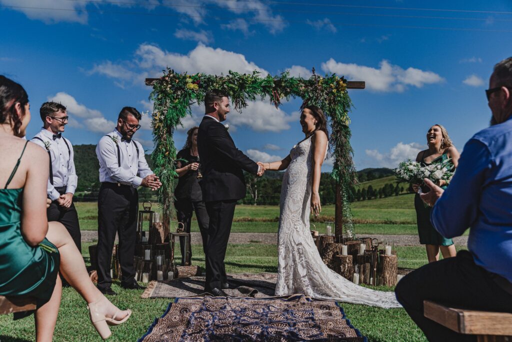 Happy Bride and groom holding hands under an eco-friendly arbour and surrounded by friends on a beautiful sunny day with blue skies