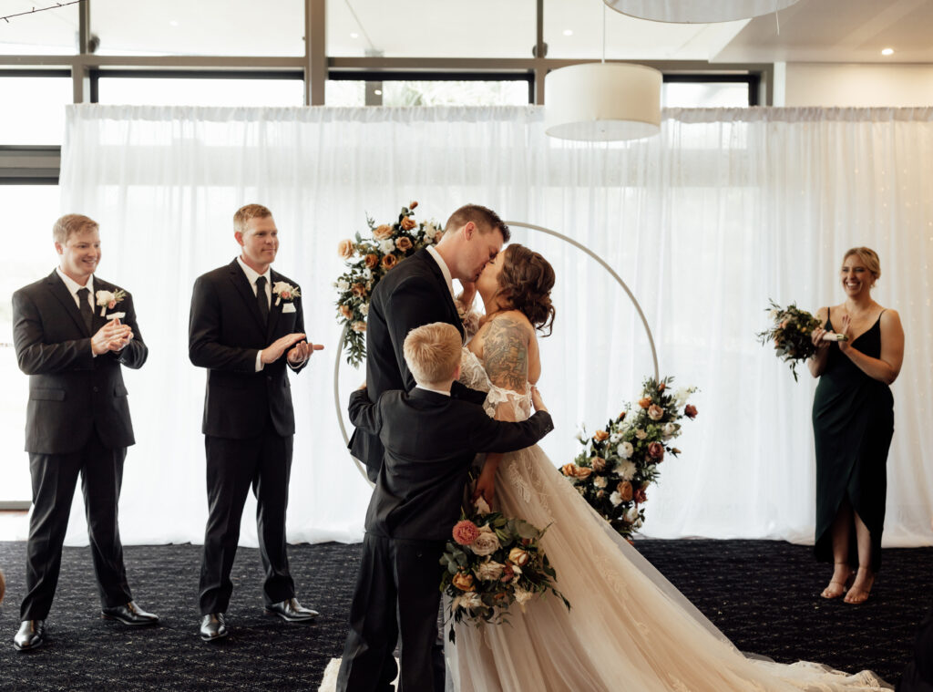 Bride and groom kissing in front of arbour as their small son hugs them both and bridal party clap and smile